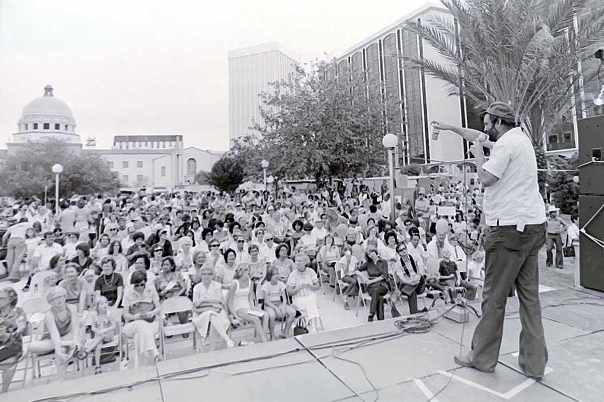 A man stands on an outdoor stage, speaking through a microphone to a large seated crowd, with a domed building in the distance. Black-and-white photo.