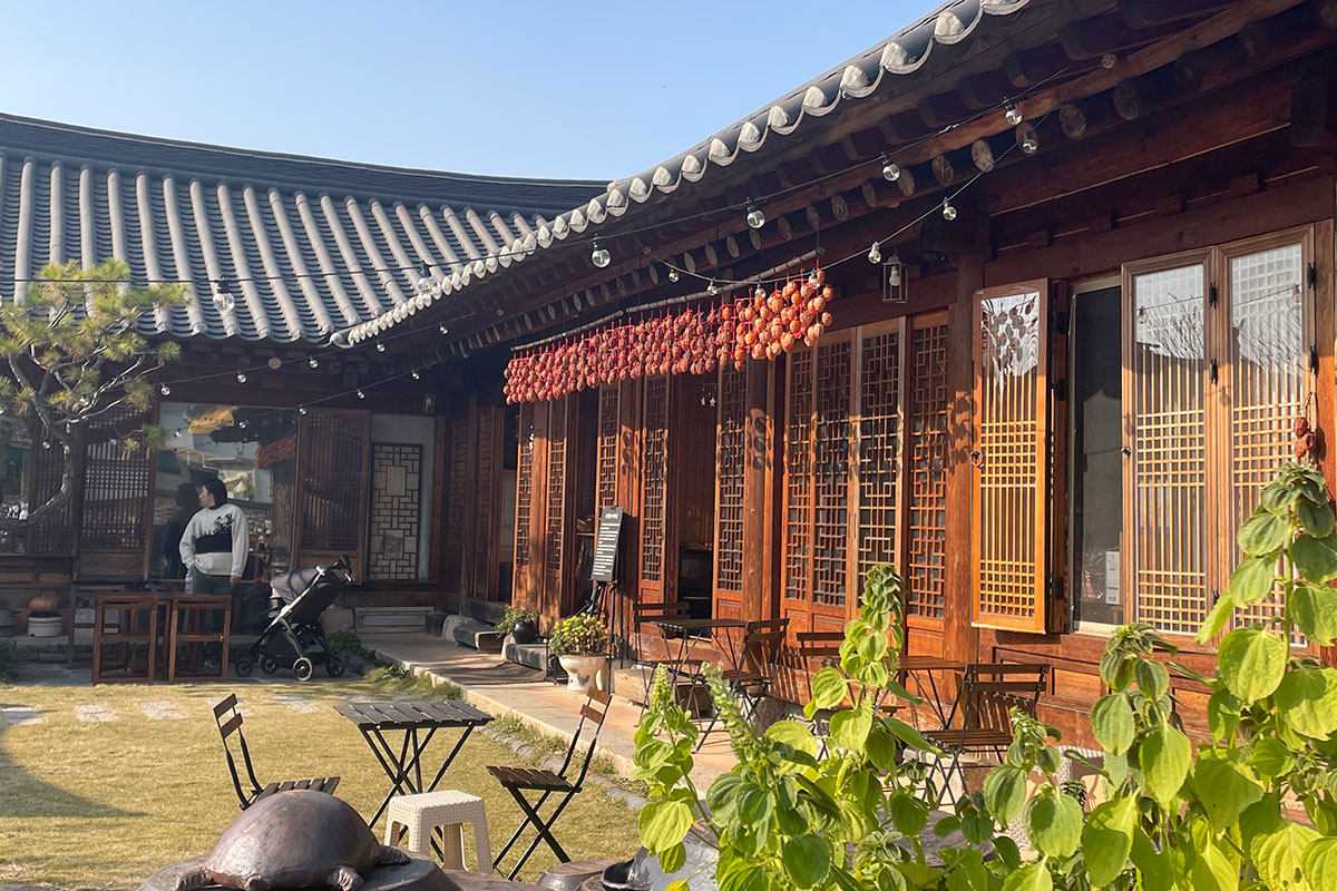 In the courtyard of a wooden home with tiled roof and carved doorways, strings of orange persimmons hang from a bamboo pole hung along an eave. 
