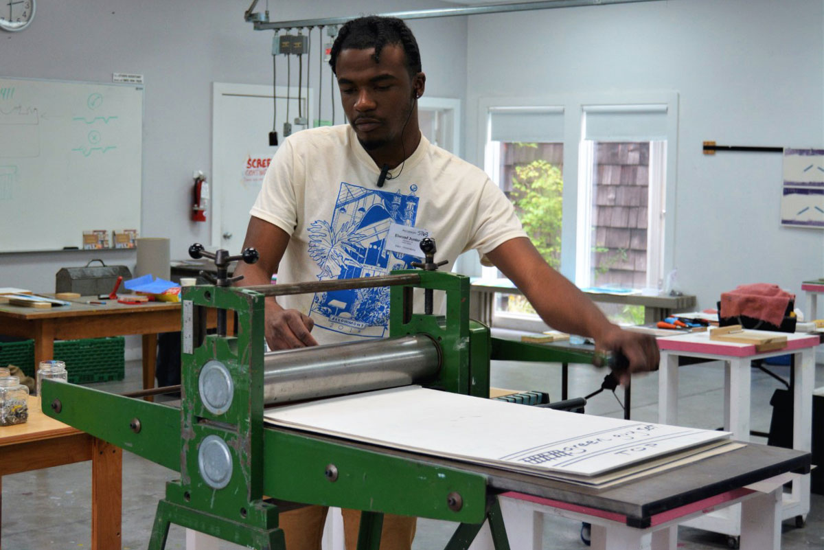 A young man pulls a crank on a green printing press in a studio work space. He wears headphones which rest on the collar of his shirt before going up to his ears.