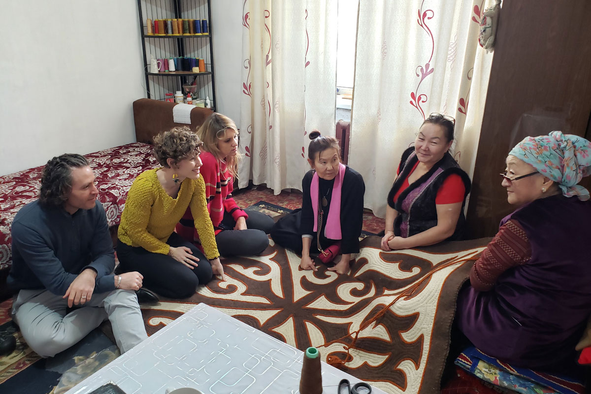 A group of people sit on the floor gathered around a traditional felt quilt made of brown and tan fabric. The woman who sewed the quilt sits above the group on a stool, explaining her process.