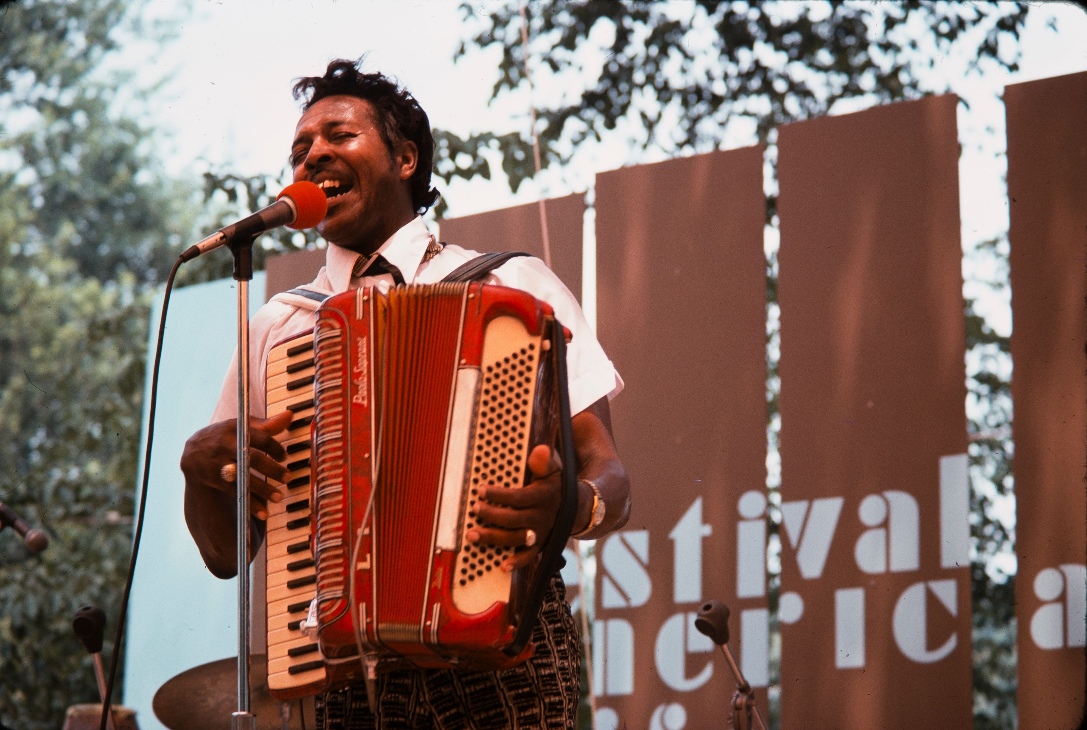 A man plays an accordion as he sings into a microphone on an outdoor stage.