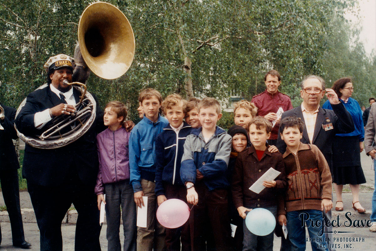 A man dressed in a black suit blows into a sousaphone, posing with a group of young kids.