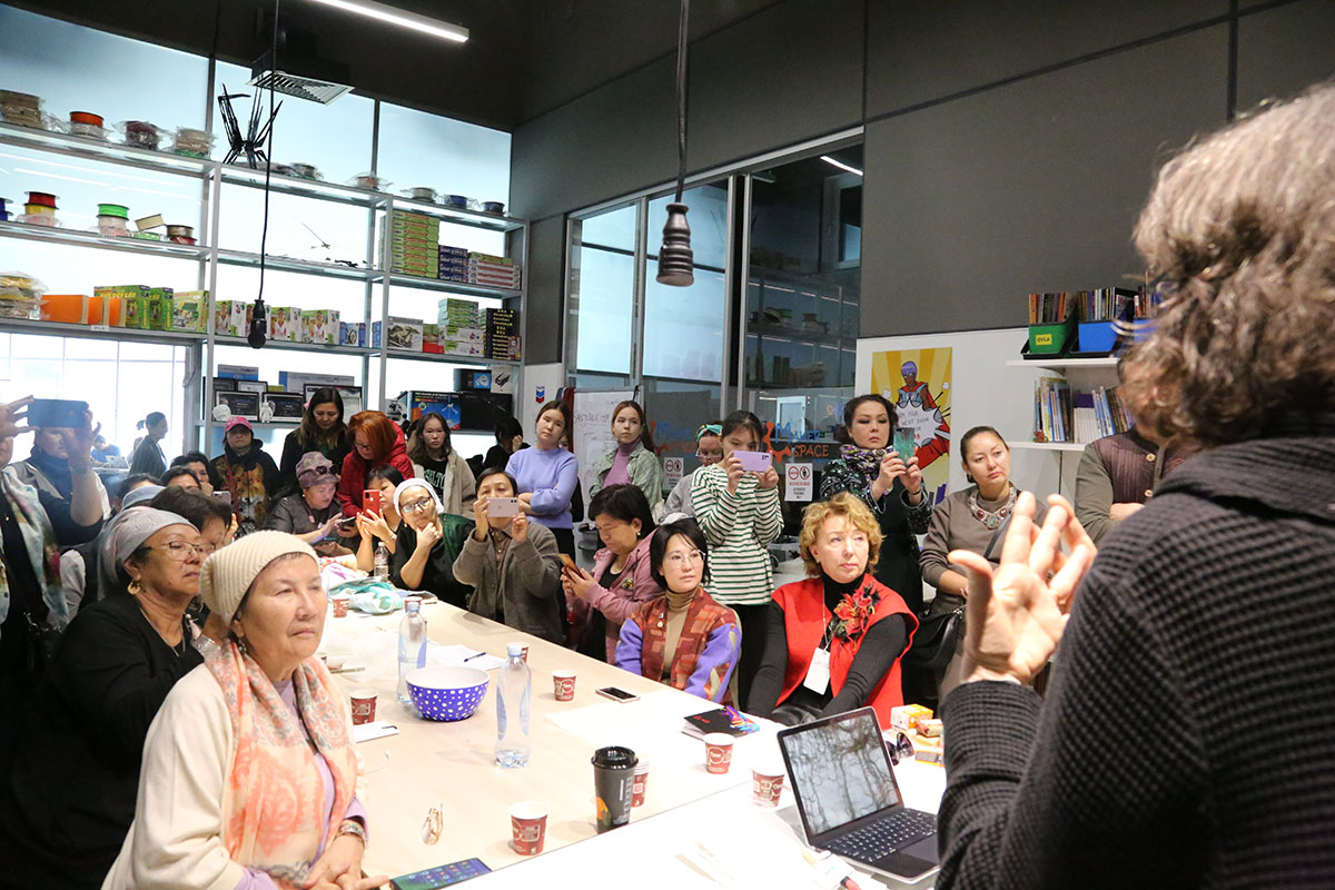 Several women crowd around a table, all facing a person at front, speaking, face turned away. Some are filming with their cellphones.