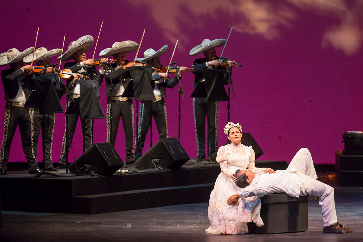 On a stage with a purple background, a man dressed in white lies on his back while a woman, also in white, puts her hands to his head and chest. Behind them, six members of a mariachi band in matching dark suits and large white sombreros, all playing violin.