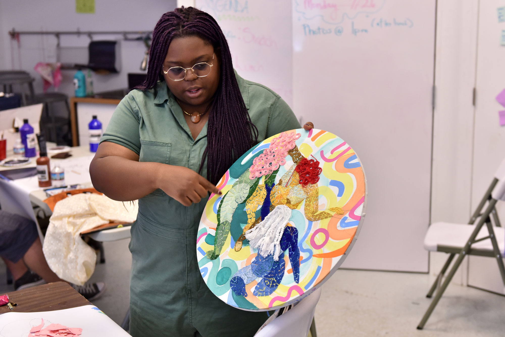 A young woman wearing a green sweater holds a circular pallet with embroidered green, yellow, red, pink, blue and white figures. She points to one of the figures while standing next to a white table and chair in front of a studio space.