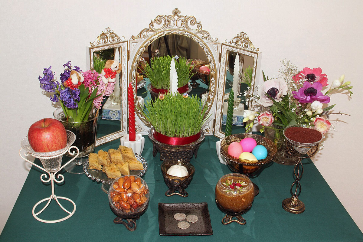 A turquoise table set with various objects: a mirror, potted grass, bouquets of flowers, an apple, dyed eggs, coins, candles, and other foods.