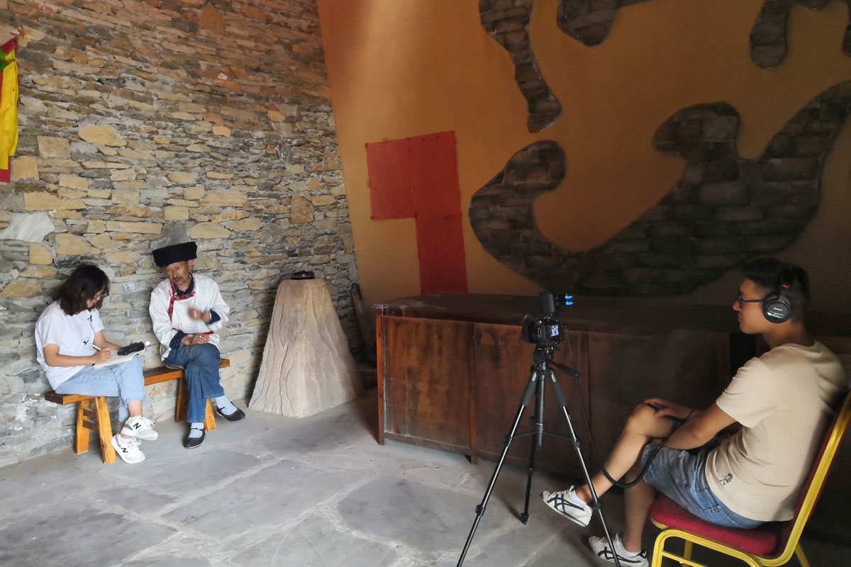 A woman holding a small audio recorder sits next to an older man wearing traditional Qiang dress in front of a stone wall. Across from them, a man stands with a video camera on a tripod.