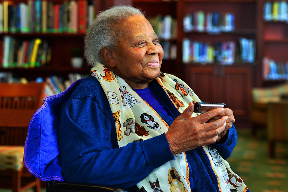 An older woman sits and smiles in a library room, holding a harmonica. Her scarf is printed with cartoon dogs.