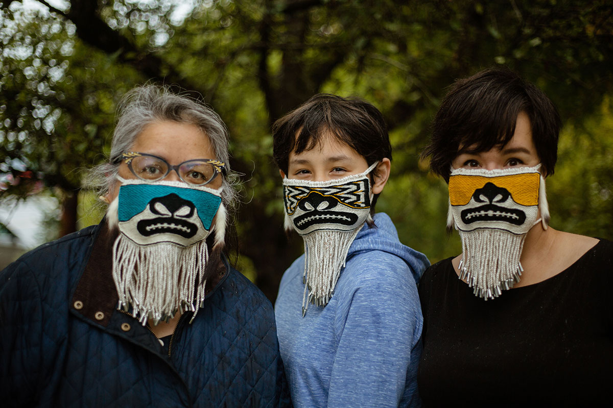 Three people, three generations of a family, pose outdoors wearing decorative beaded face masks.