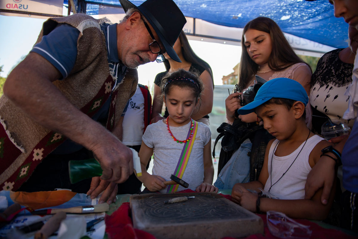 An older man stands to the right of the frame holding a spray bottle over a stone he is carving. To his right stand a small crowd of children watching him work.