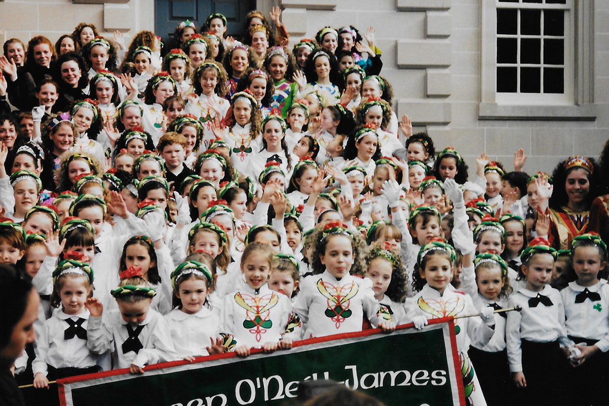 Posed group photo of dozens of young kinds on the steps to a building. they are wearing matching white and green outfits.