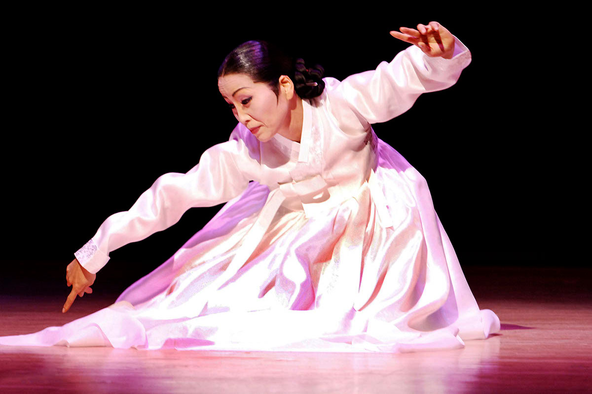 A woman in a long-sleeved pink gown crouches on a stage in a dance, with a spotlight on her.