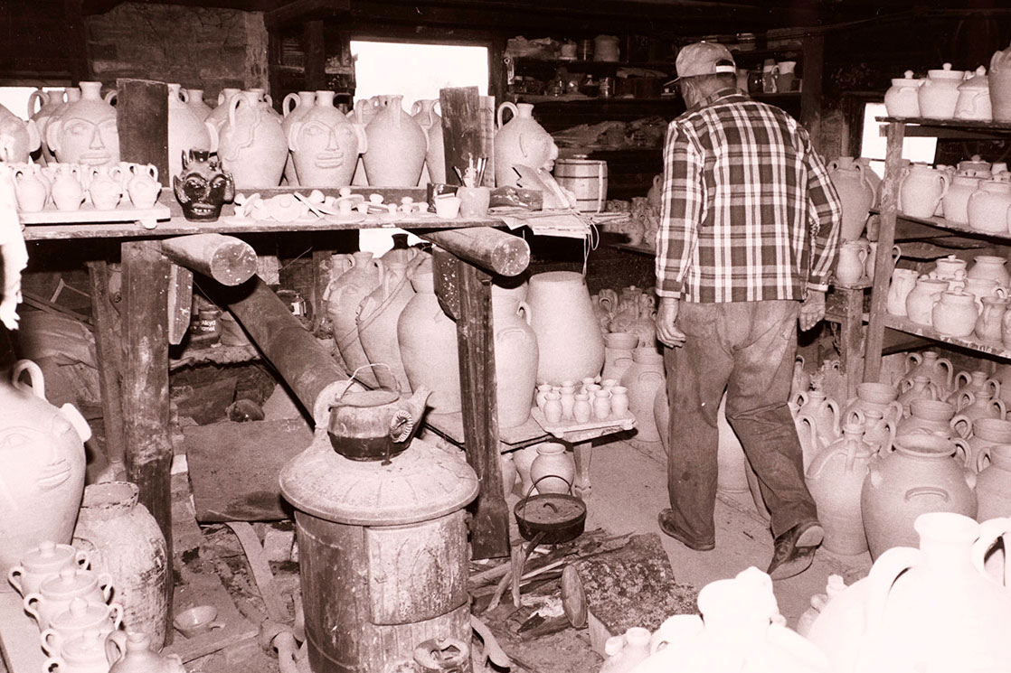 A man, facing away, walks through a workshop room. Every flat surface, including the floor, shelves, and tabletops, contains ceramic jugs in progress. Some of them have humanoid faces on the sides.