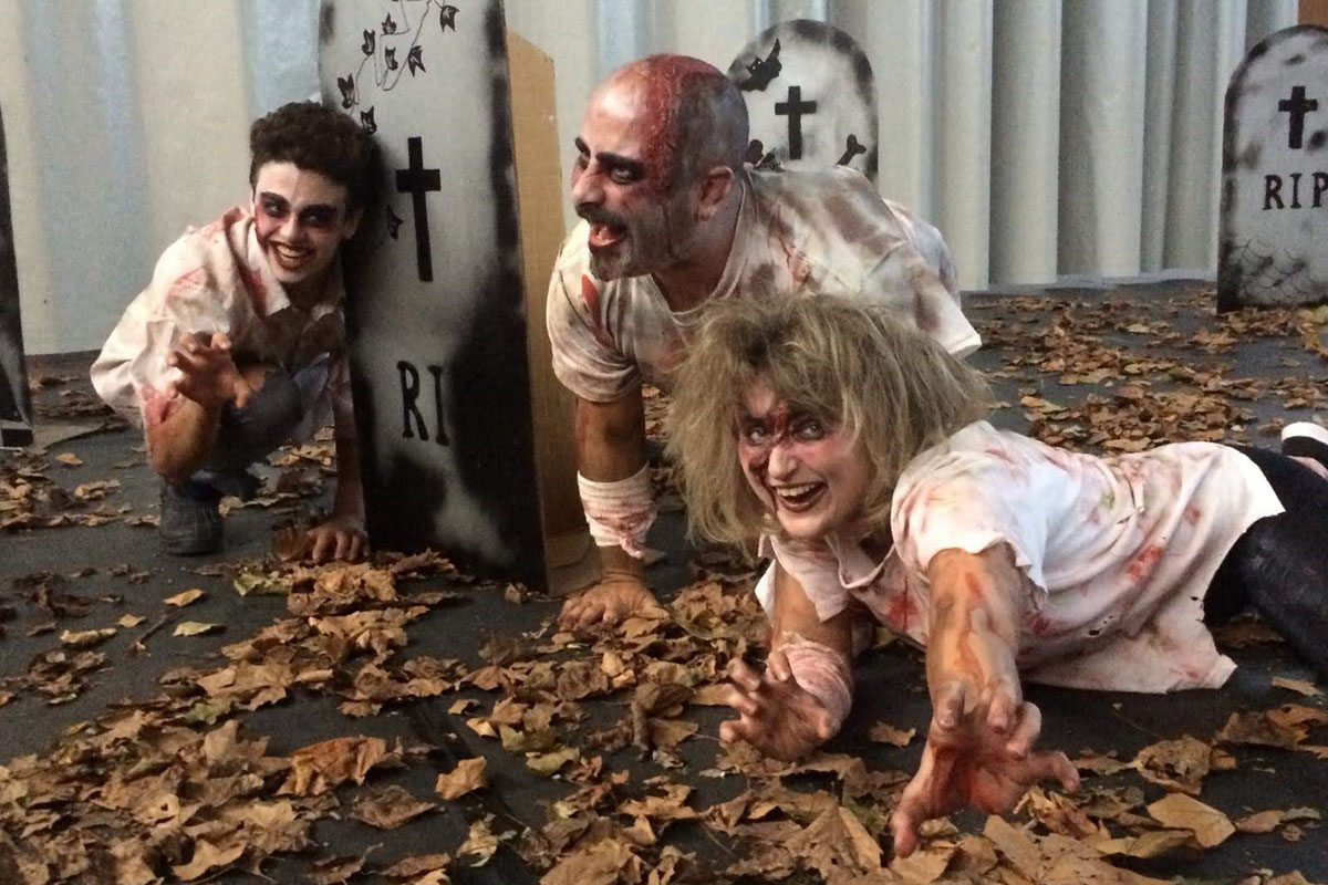 Three people act as blood-splattered zombies in a street filled with dead leaves and cardboard gravestones.
