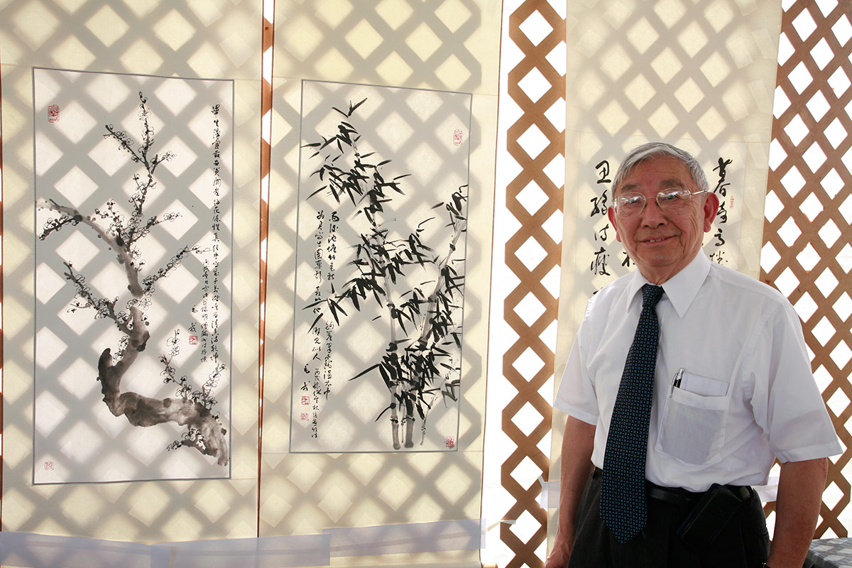 A elder man with short gray hair, glasses, and a blue tie with white button-up shirt poses besides hanging vertical paintings, depicting plum blossoms and bamboo with Chinese characters.