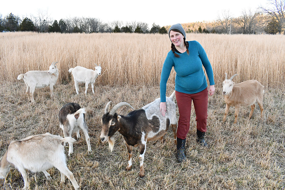 A woman pets one of six goats on a farm in winter, with bare trees and a field of golden grain behind her.