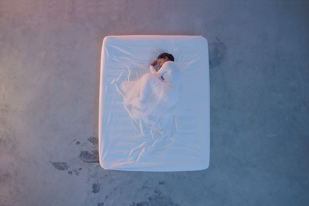 From above, a woman dressed in white, curled in fetal position, under a white sheet on a white mattress on a concrete floor.