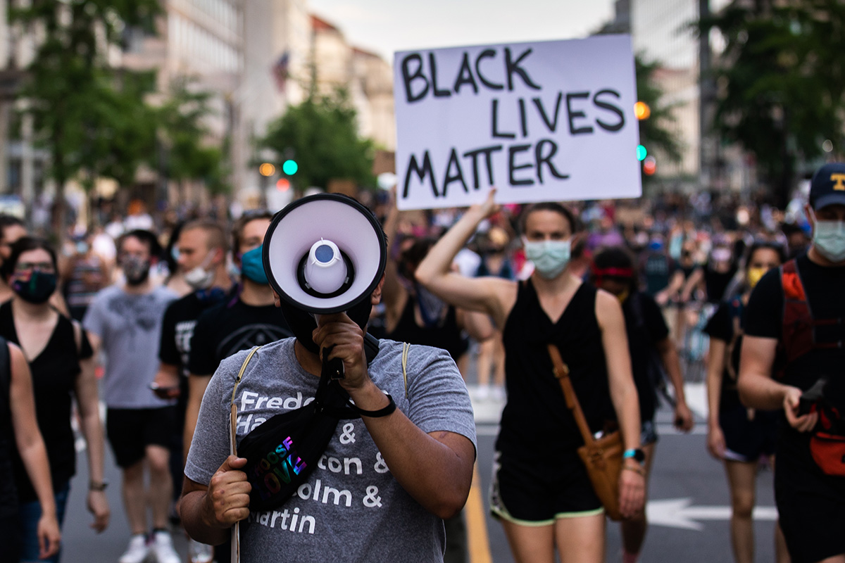 Countless people marching the street. In the foreground, a man with his face obstructed by a megaphone. Behind him, a woman holding up a sign reading BLACK LIVES MATTER. Behind them, out of focus, many people marching toward the camera, many in face masks.