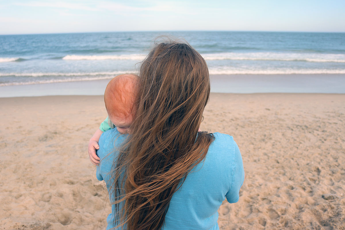 A woman standing on the beach faces the ocean, holding a small red-headed baby. Both their faces are hidden.