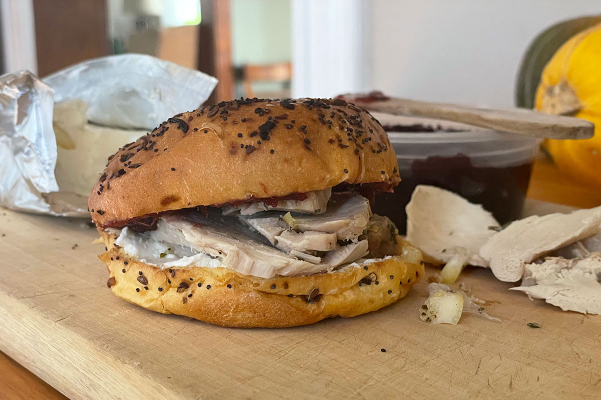 Close-up of a sandwich on a wooden cutting board: sliced turkey, white cream cheese, and dark red cranberry sauce on a seeded bun.
