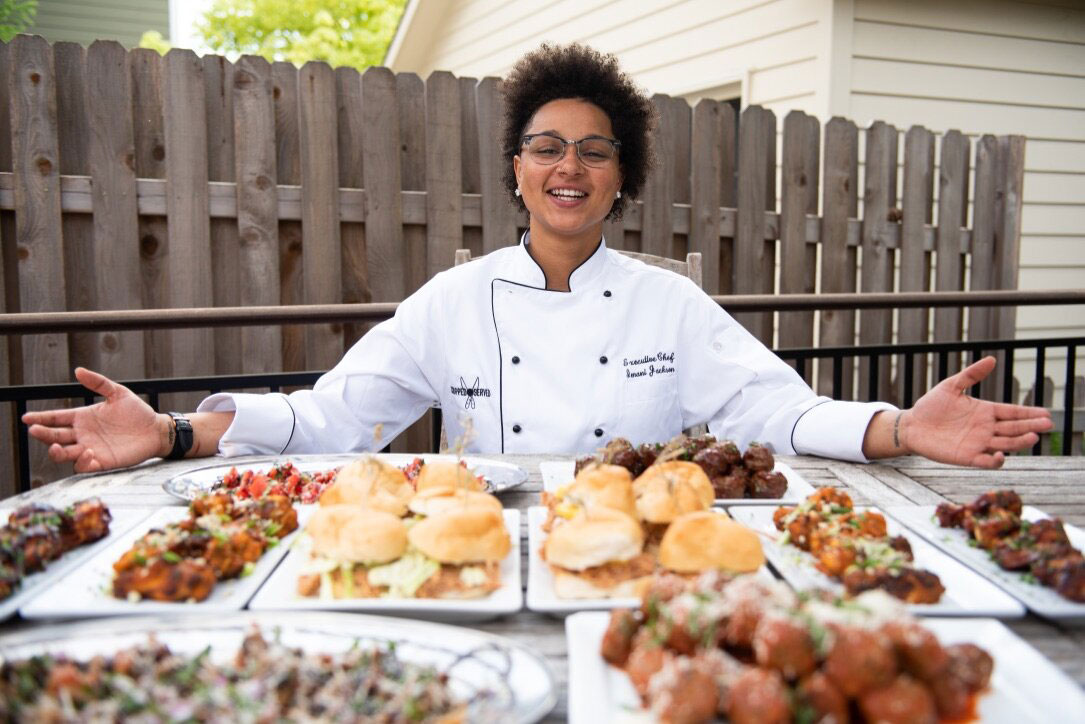 A Black woman in a white chef's coat opens her arms toward a table full of food.