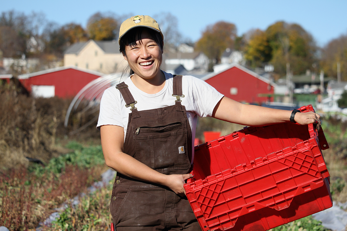 A young Asian woman in white T-shirt and brown overalls holds a red plastic container, smiling at the camera, with a farm behind her.