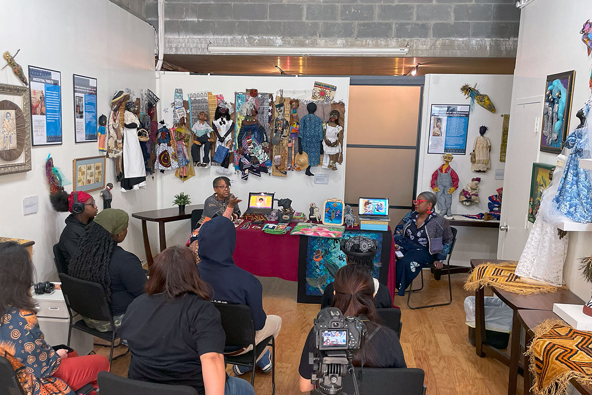In a narrow art gallery room, young people sit in chairs facing two women at a table full of textile art. A camera on a tripod records them in the foreground.