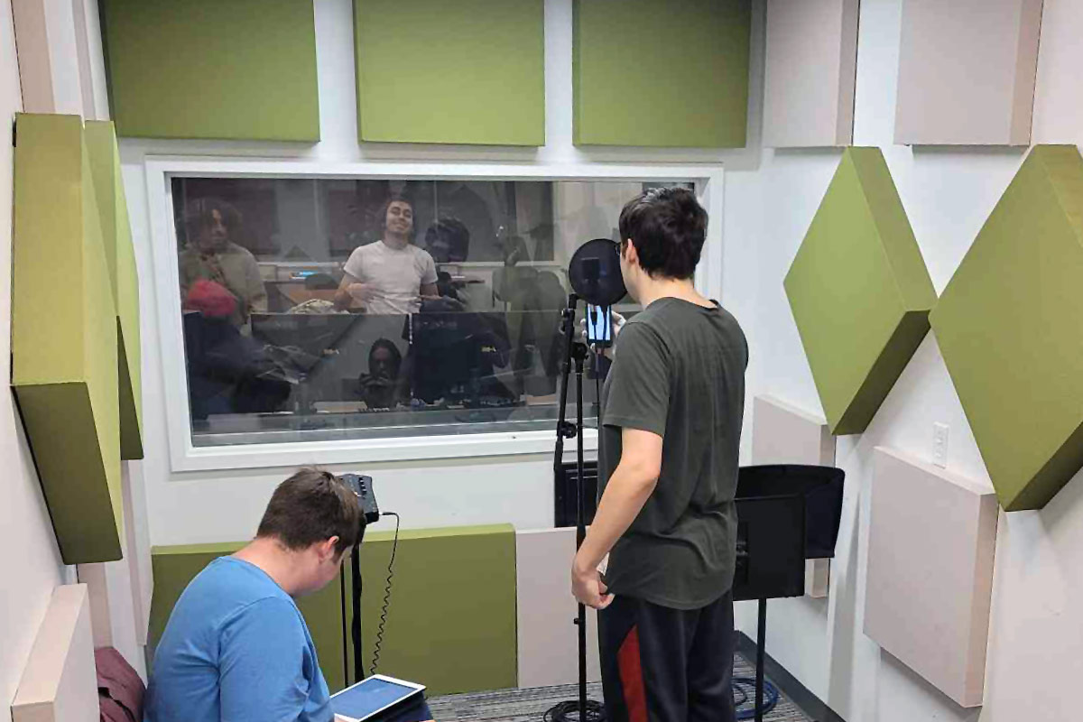 In a small room lined with green and beige-colored square panels on the walls, a person stands in front of a microphone, facing others through a control window.