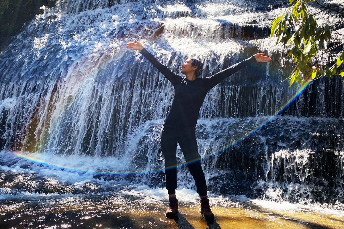A woman sticks on a smooth rock surface in front of a wide waterfall. Its spray creates two arcs of rainbows that frame her, with arms outstretched.
