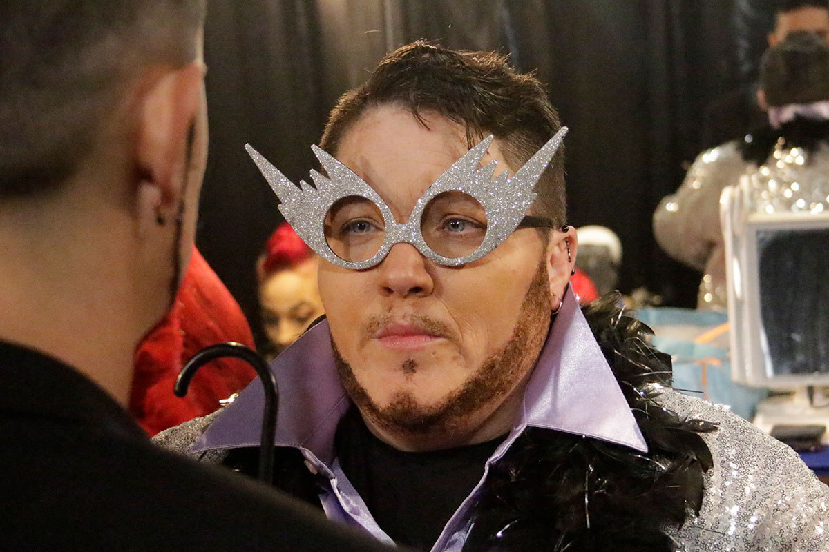 A person with glittery silver coat with oversized lavender collar, black feather boa, prop glasses - no lenses but silver frames that resemble comets with jagged trails behind them - and a thin faux beard faces another person in a mirrored dressing room.