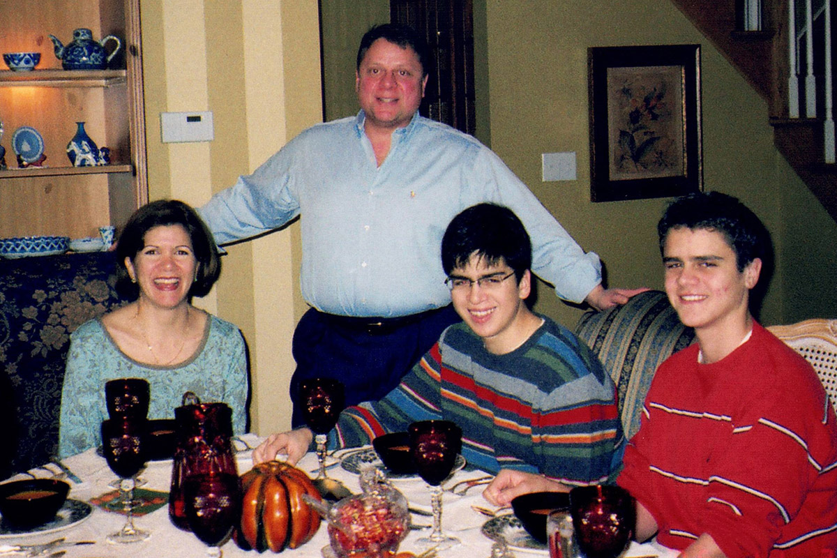 A family of four at an elaborately set dinner table: mom and two brothers seated, and dad standing between them.