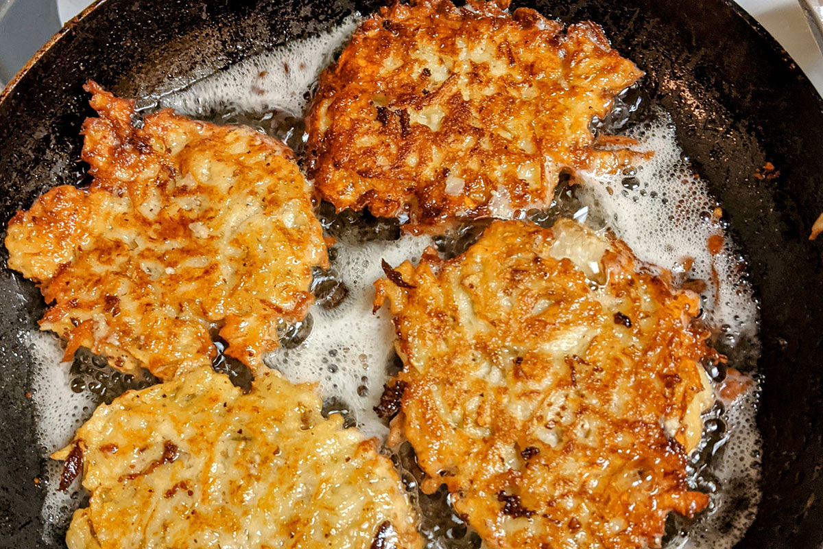 Four golden brown, shredded potato pancakes frying in a cast iron pan.