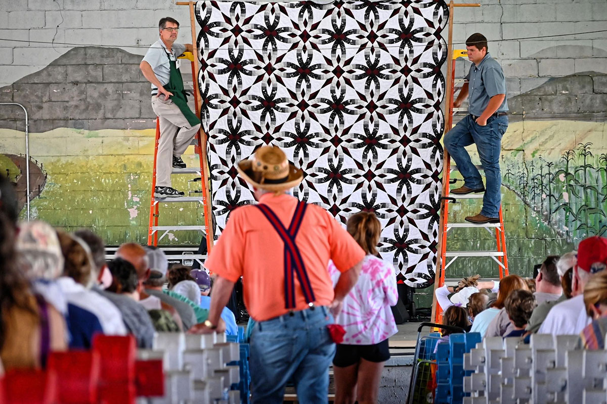In front of a seated and standing crowd, two men on ladders show off a large hanging quilt with black and white geometric design.