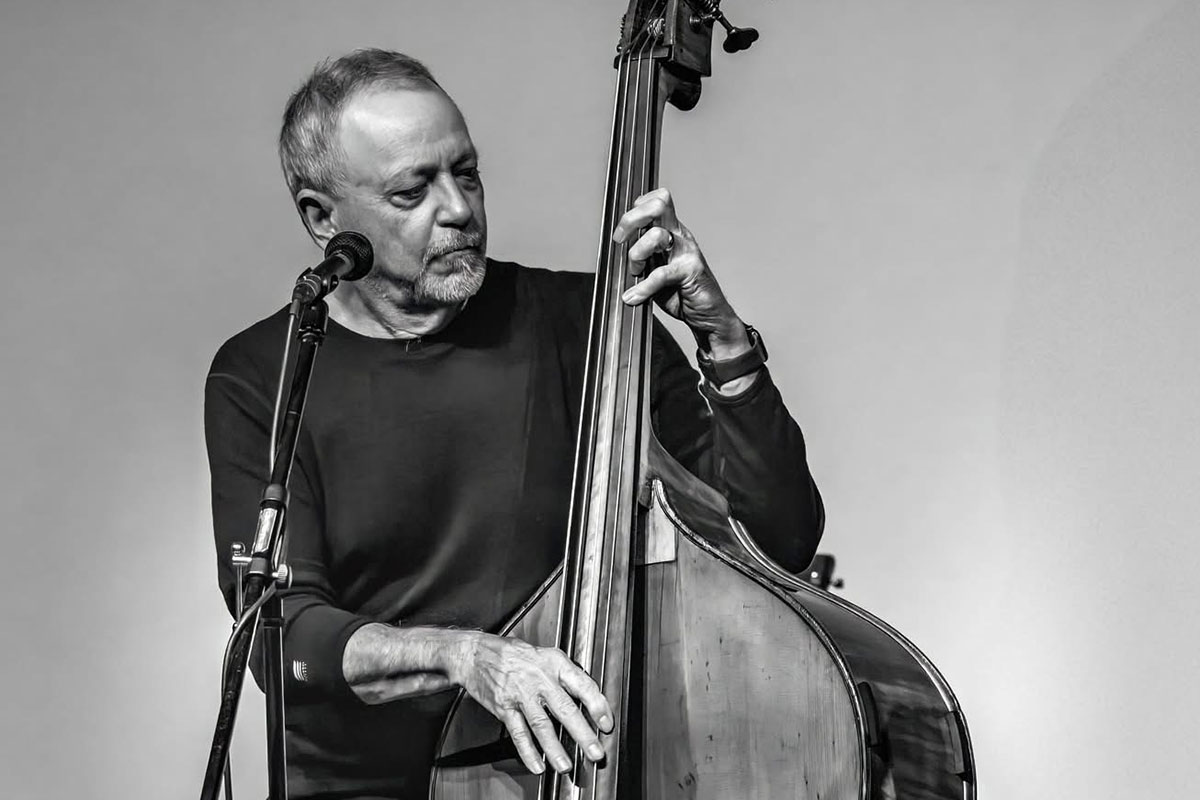 A man with short gray hair and black shirt plays upright bass on stage. Black-and-white photo.