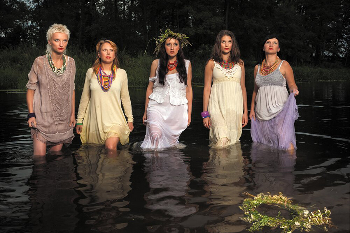 Five women in flowy, pastel-colored dresses stand knee-deep in water at dusk. In the foreground, a wreath floats on the surface of the water.
