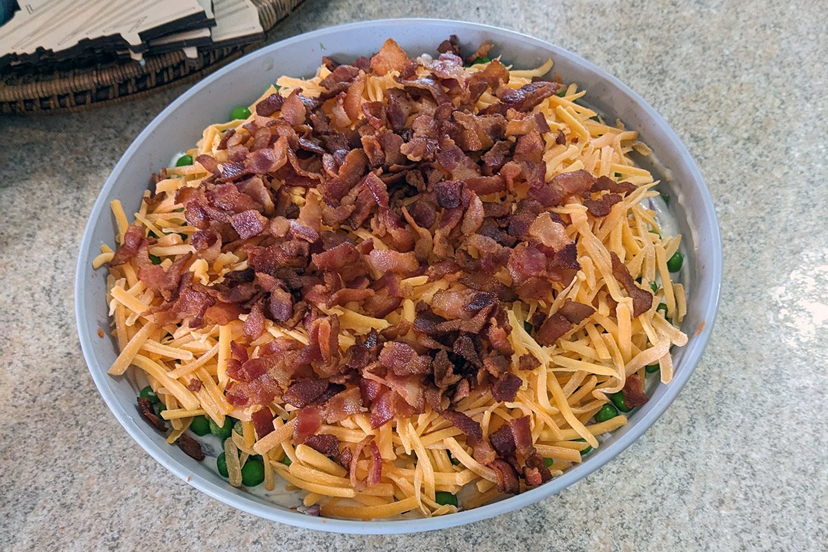 In a circular bowl, red bacon bits and orange cheddar cheese top a salad. Green peas and white dressing peak through from below.
