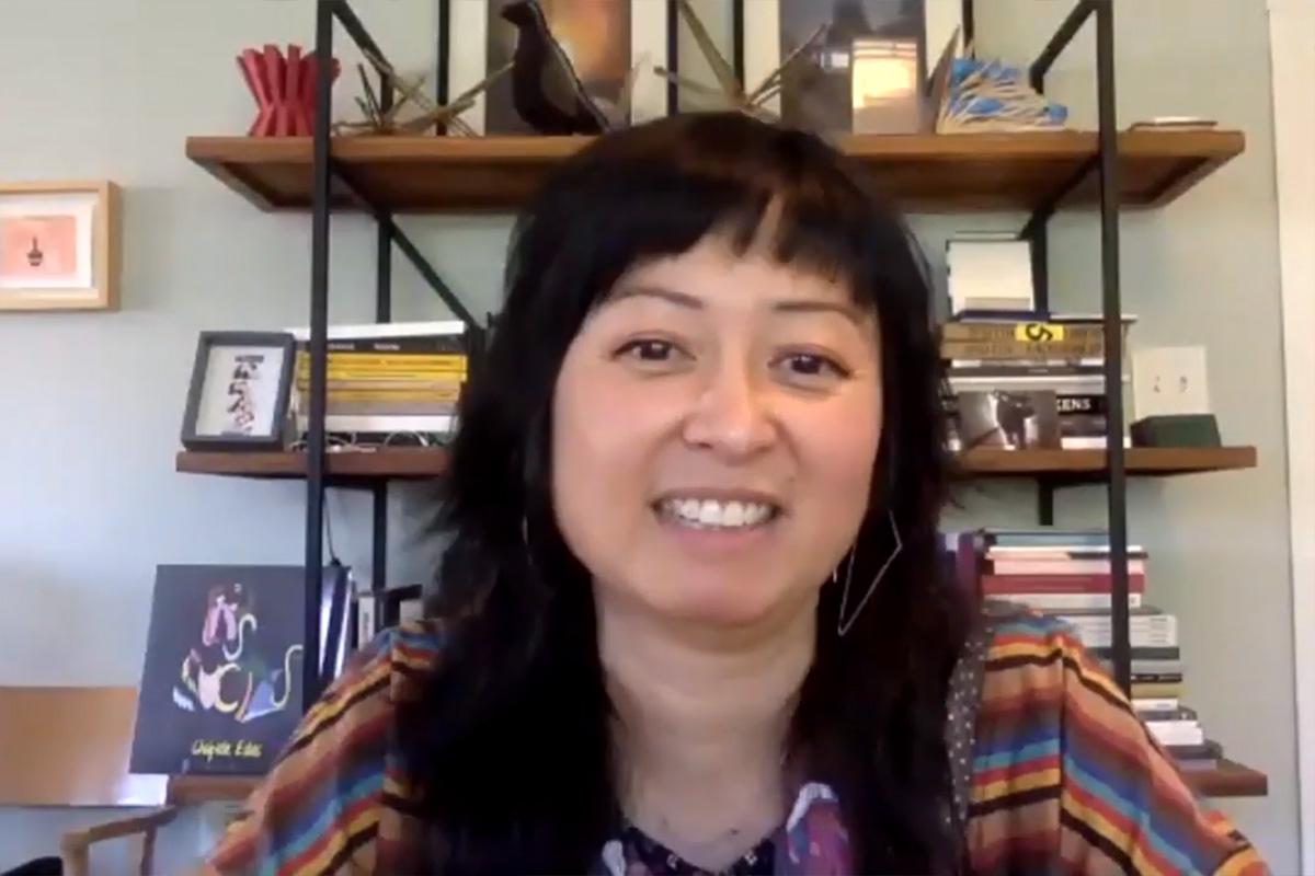 Stephanie Syjuco smiles at the camera in a screenshot of a virtual conversation. She wears a colorful striped top and angular earrings, with a shelf full of books and art behind her.