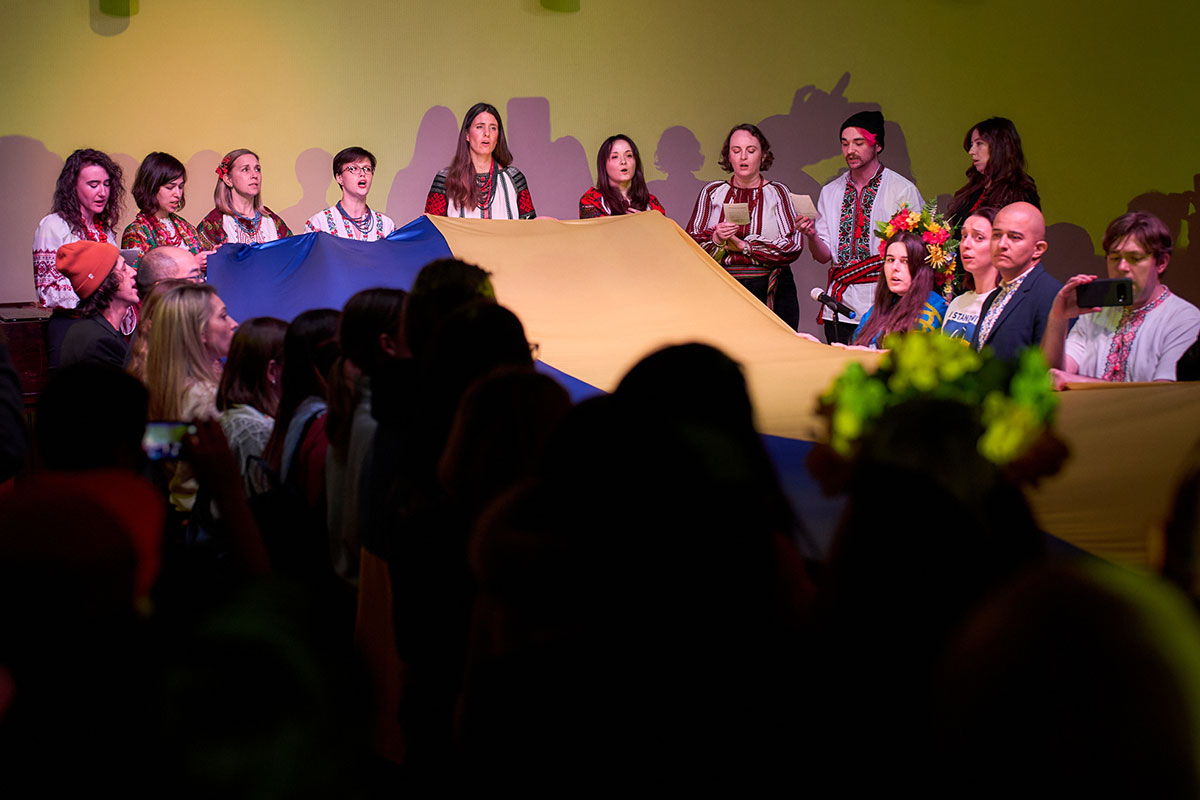 A crowd of people holds up a gigantic Ukrainian flag, half yellow, half blue. On the far side of the flag, a group wearing traditional embroidered tops sing.