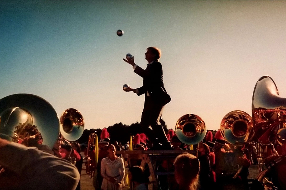 At dusk, silhouetted figure of a man juggling, surrounded by the glinting bells of sousaphones and marching band members in uniform.