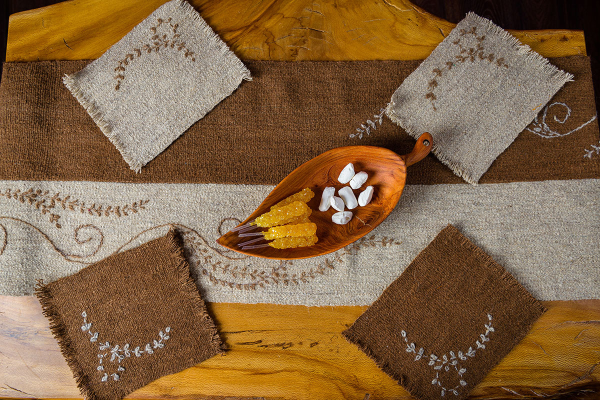 From overhead, a wooden table set with a wool runner in brown and cream and four matching wool coasters, all embroidered with a vine pattern. In the center of the table, a wooden leaf-shaped dish holds yellow rock candy and white tablets.