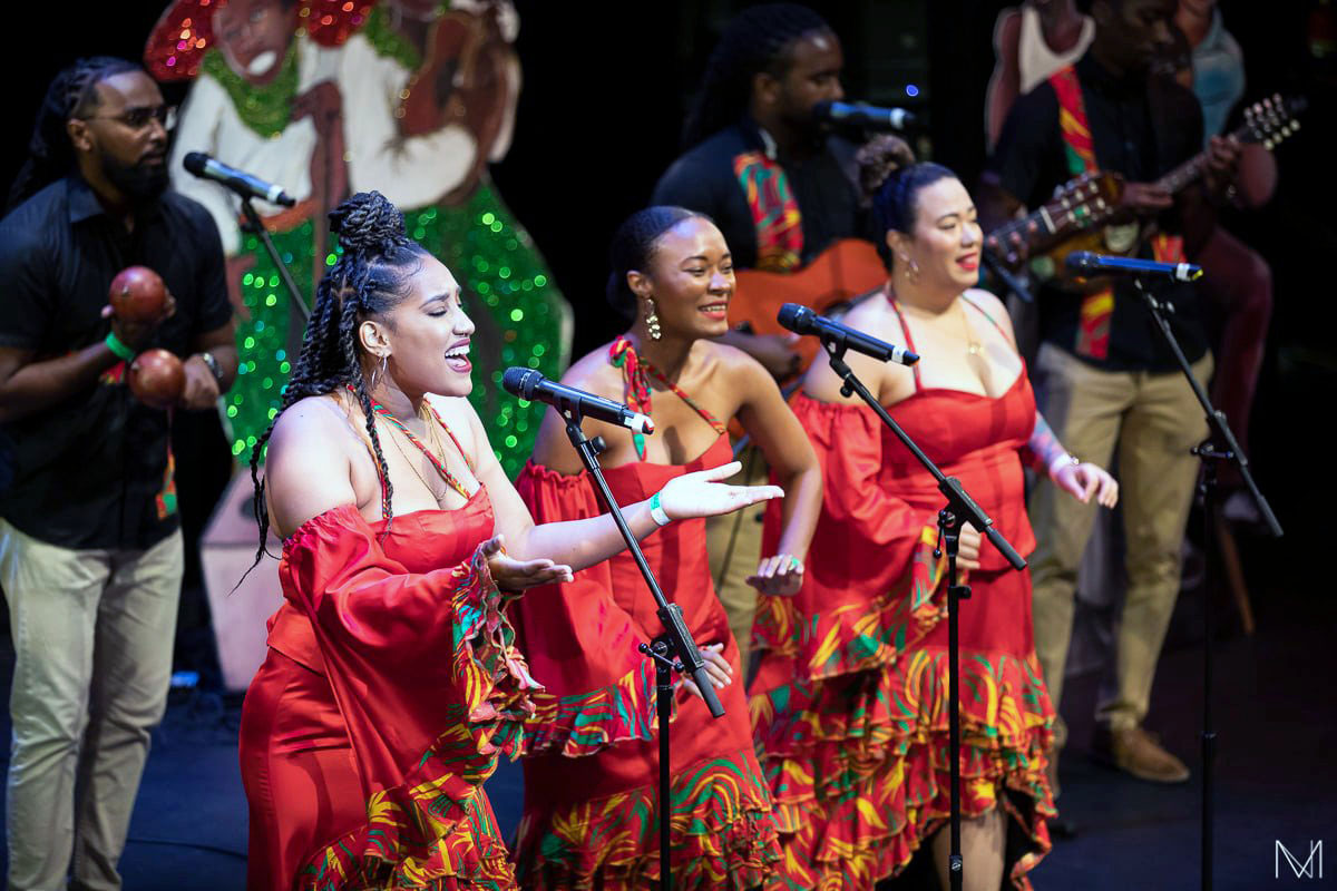 Three women singers in matching red dresses perform on stage, with male musicians and Christmas decorations on stage behind them.