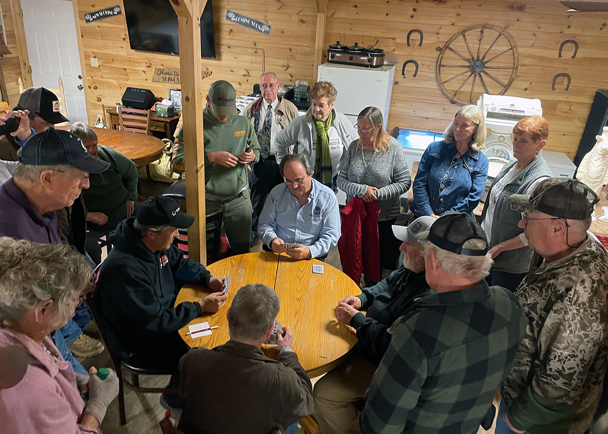 Four men sit around a round wooden table playing cards, while a crowd of others surround them.