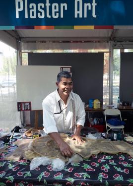 Aguilar, wearing a white embroidered shirt, smiles from his workspace under a tent and a banner that reads Plaster Art.