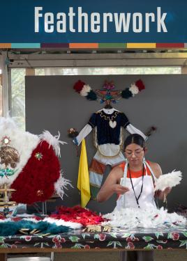 Ramirez, wearing an orange tank top and white apron, handles a bouquet of white feathers in her workspace under a tent and a banner that reads Featherwork.