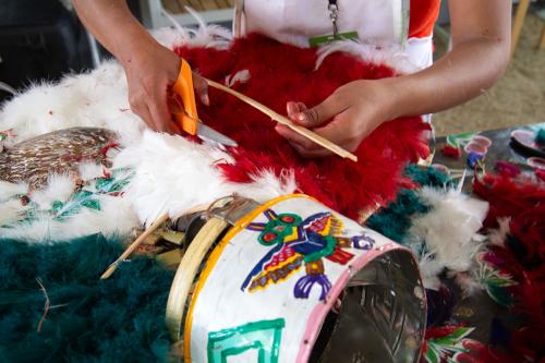 Close-up on a woman's hands as she uses scissors to trim white and red feathers on a decorative headdress. The crown of the piece features a stylized image of a bird. 