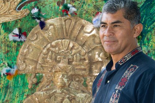 A man in a black embroidered dress shirt looks into the distance, standing beside a plaster relief work of a human figure wearing a semicircular headdress on a painted green background.