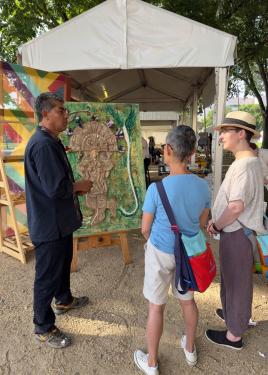 The man, dressed in all black, stands behind the green plaster relief work, set on an easel outdoors, as he talks with two festival visitors.