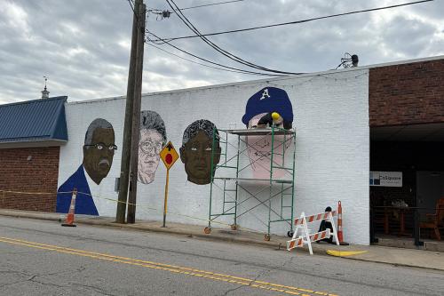 Seen from across the street, a man crouches on scaffolding, painting the upper half of a mural on the side of a  building under gray skies.