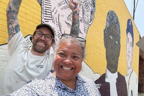 A man and woman take a selfie in front of the mural with a  yellow background. They both smile broadly, while the man raises both his tatooed arms in joy.