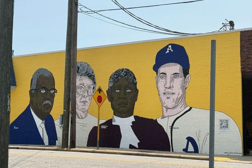 Ground-to-roof mural on the side of a brick building, showing four people on a yellow background. Left to right, the figures are an elder Black man with blue suit jacket and gray moustache, elder white woman with short gray hair and glasses, elder Black woman with maroon coat, and a white man with blue ball cap and white baseball jersey.